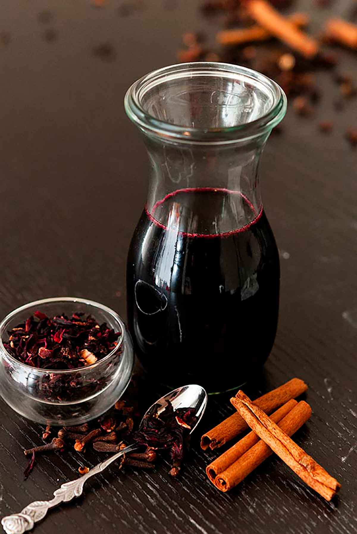 A bottle of spiced hibiscus syrup on a table surrounded by a few cinnamon sticks and a small bowl of hibiscus flowers.