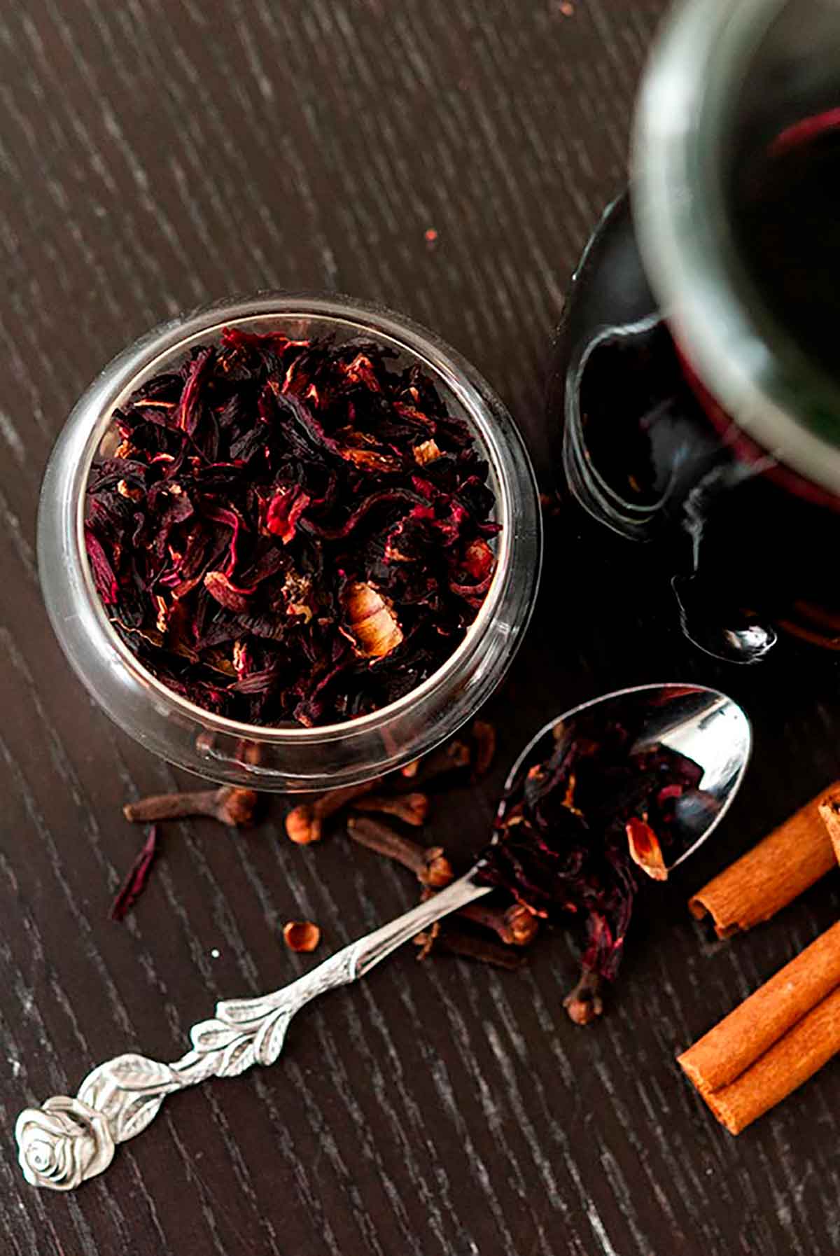 Dry hibiscus flowers in a small bowl next to a small spoon with a few scattered cloves, flowers and cinnamon sticks.