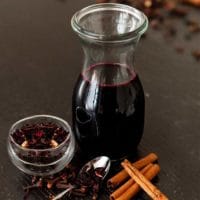 A bottle of spiced hibiscus syrup on a table surrounded by a few cinnamon sticks and a small bowl of hibiscus flowers.