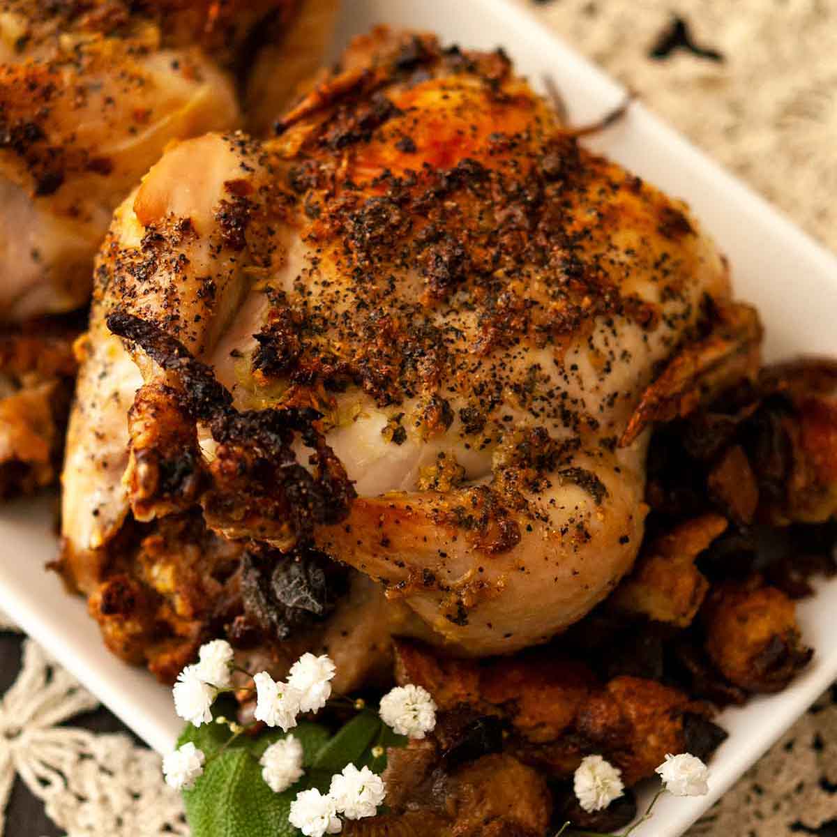 A cooked Cornish game hen on a plate, on top of a lace doily, beside stuffing, a few flowers and 2 sage leaves.