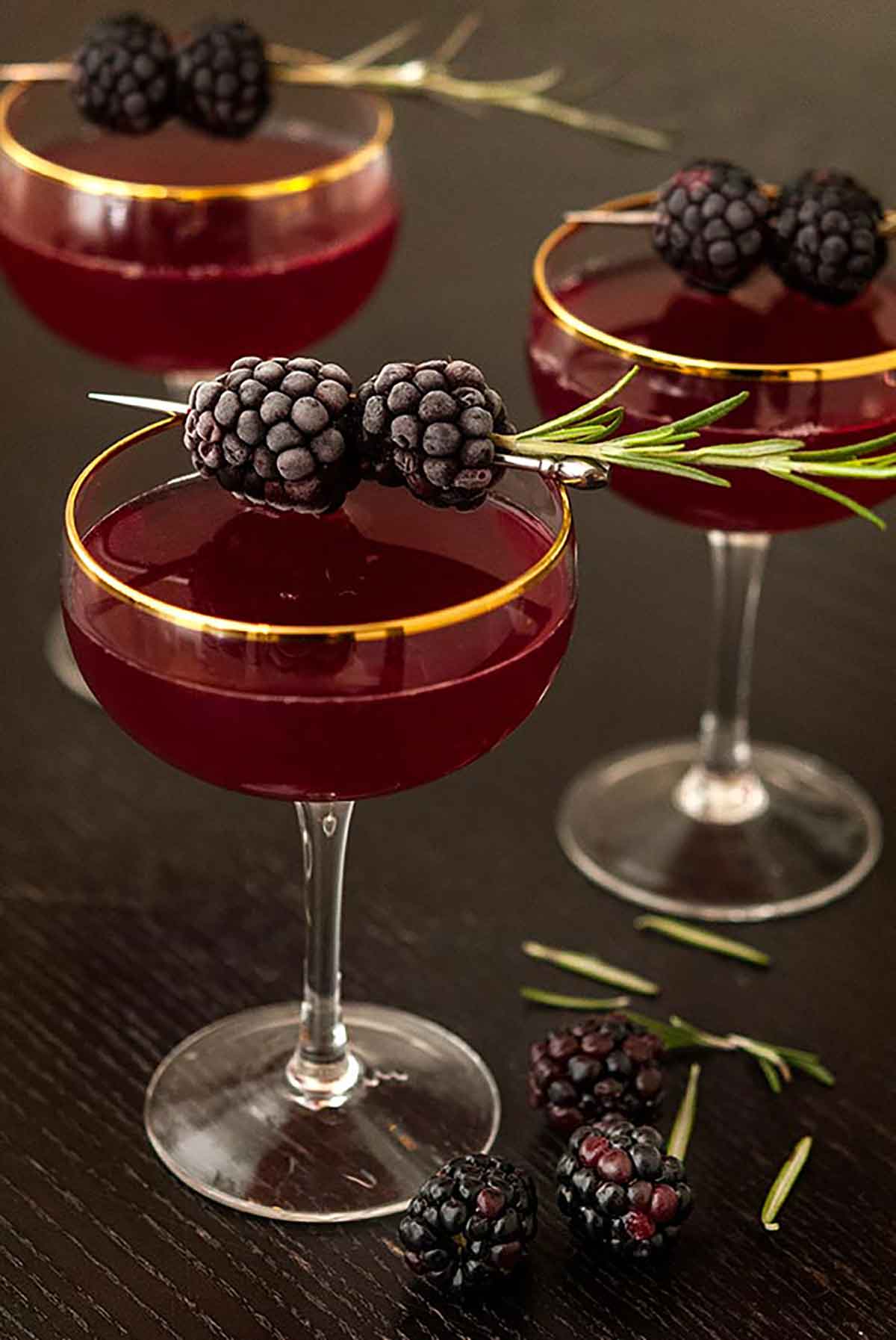 3 cocktails on a table surrounded by a few scattered blackberries, and garnished with frosted blackberries and rosemary.