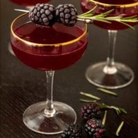 A cocktail surrounded by a few blackberries, garnished with 2 frosted blackberries and a cocktail in the background.