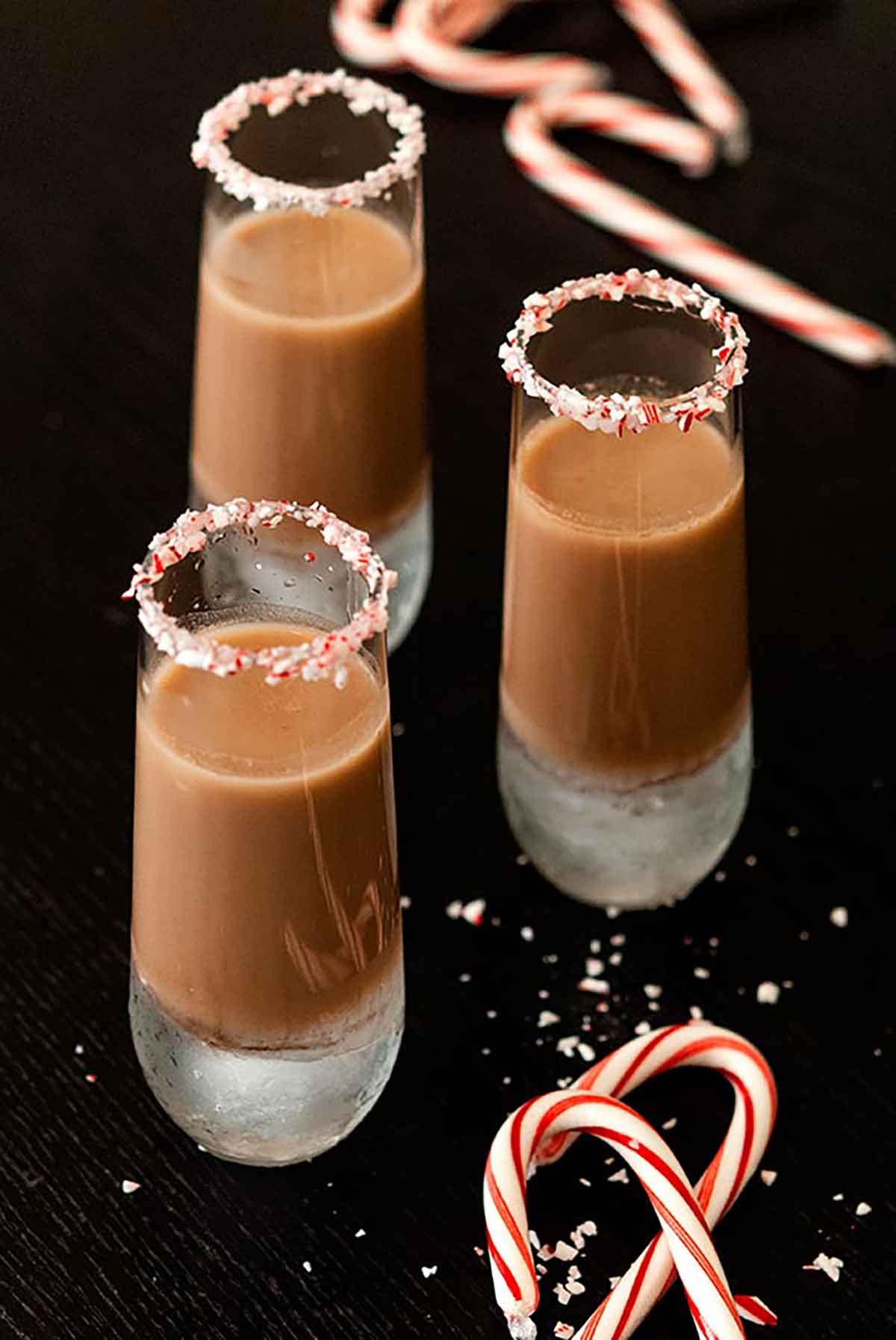 3 chocolate cocktails, rimed with crushed candy cane on a black table with a few scattered candy canes.