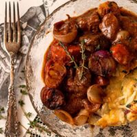 A bowl, beside an antique fork, of beef bourguignon on a table, covered in images of 1800’s fashions.