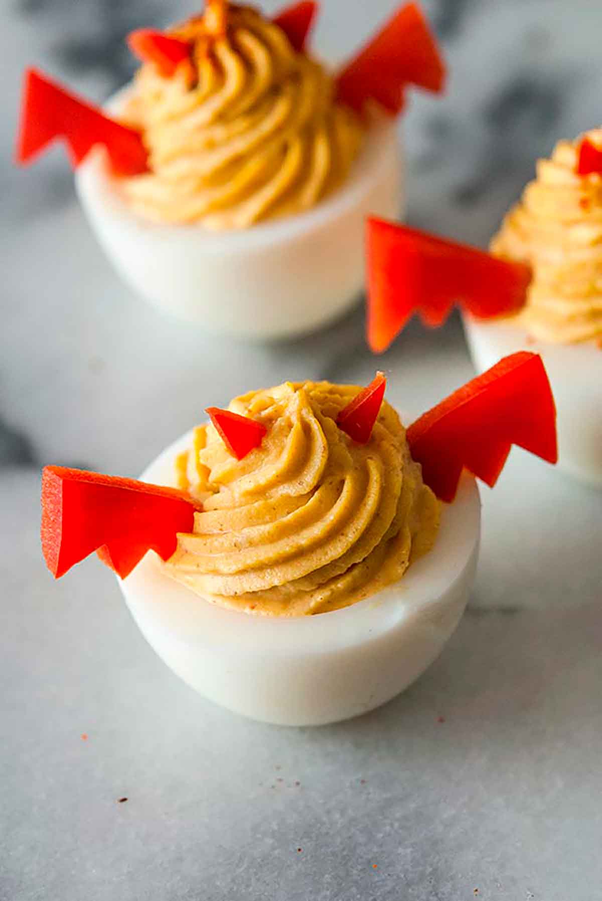 3 deviled eggs garnished with horns and wings made of red pepper on a marble board.