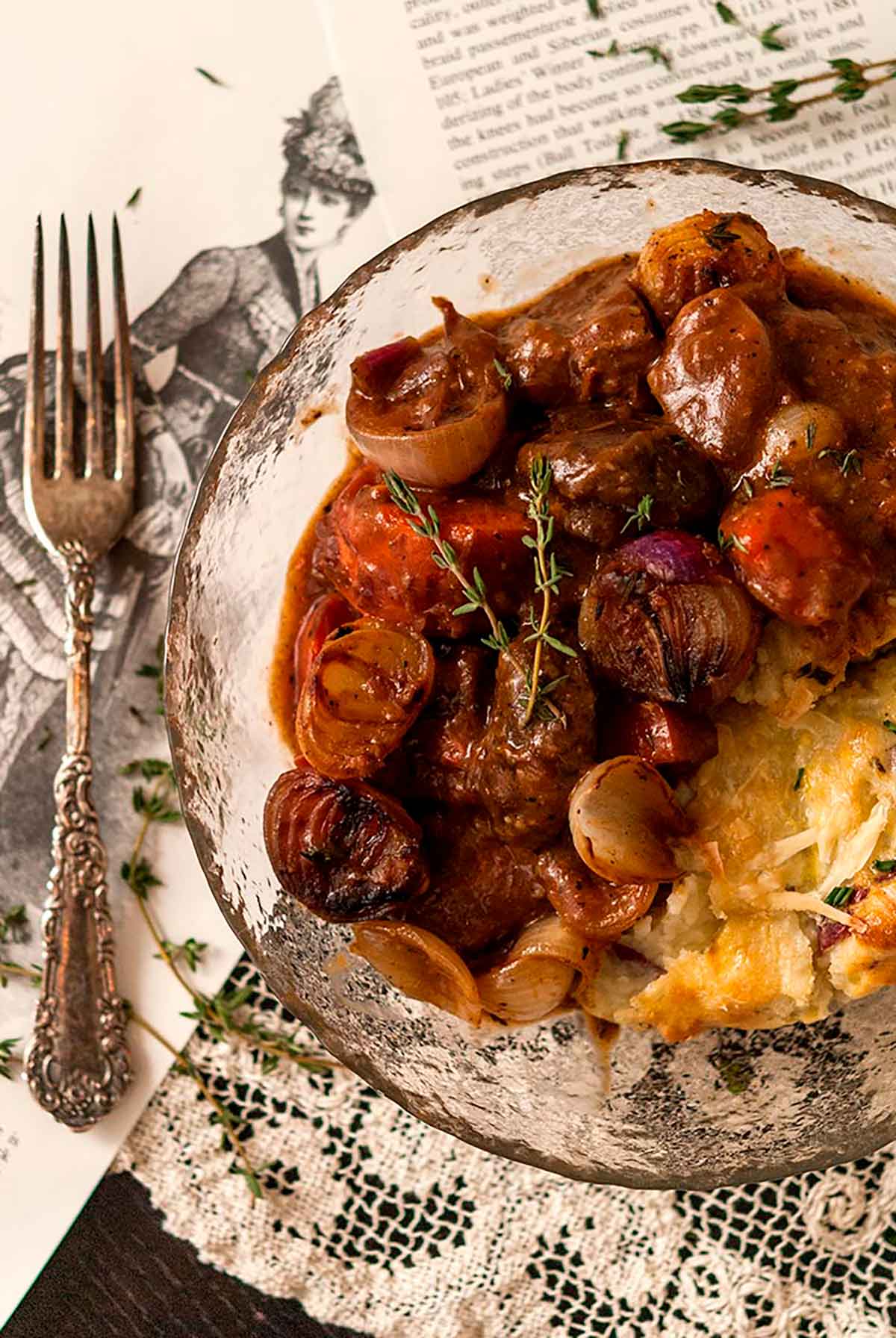 A bowl, beside an antique fork, of beef bourguignon on a table, covered in images of 1800’s fashions.