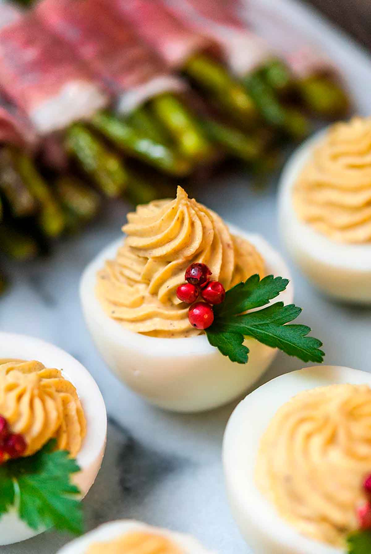A deviled egg garnished with pink peppercorns and parsley, surrounded by other deviled eggs and asparagus.
