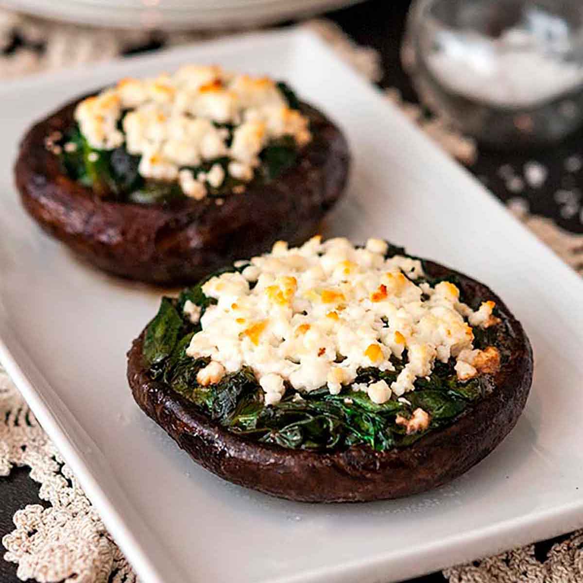 2 spinach and feta-stuffed mushroom caps on a plate on a table, lined with lace with 2 stacked plates in the background.