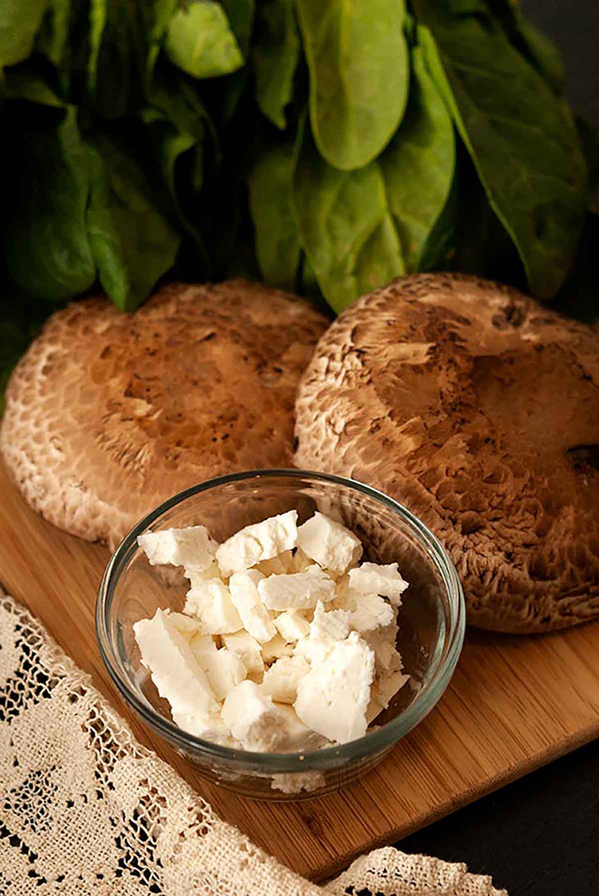 2 mushroom caps, a bowl of feta and a bunch of spinach leaves on a cutting board.