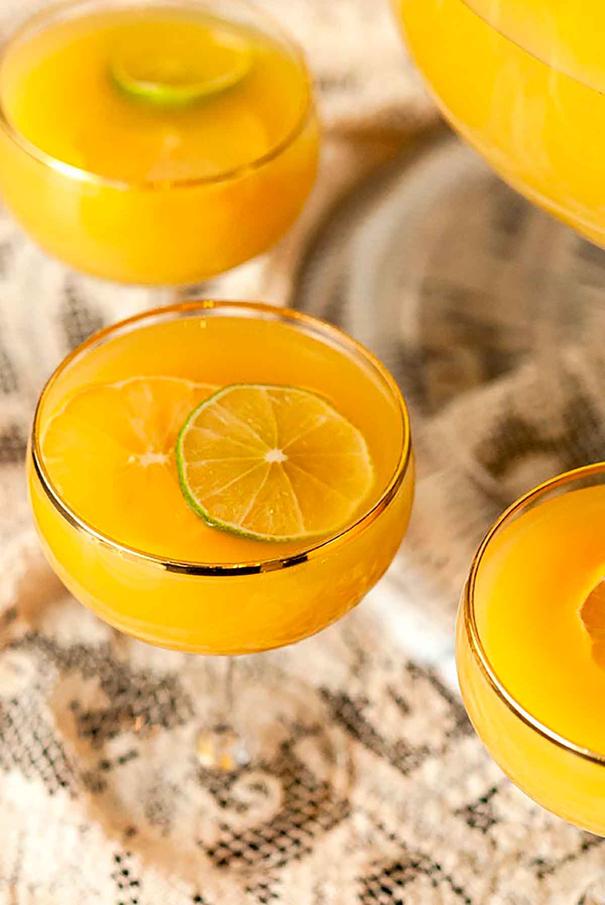 3 glasses of pineapple punch, garnished with limes on a lace table cloth beside a punch bowl.