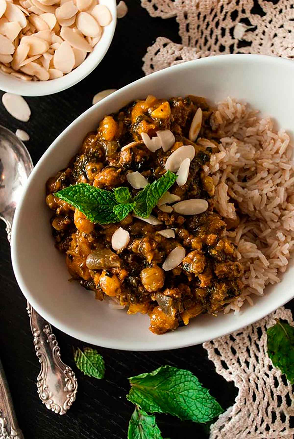 A bowl of turkey curry on rice on a lace doily beside a bowl of sliced almonds and a spoon.