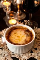 A bowl of French onion soup on a small lace doily in front of a candle, candle stick and glass of red wine.