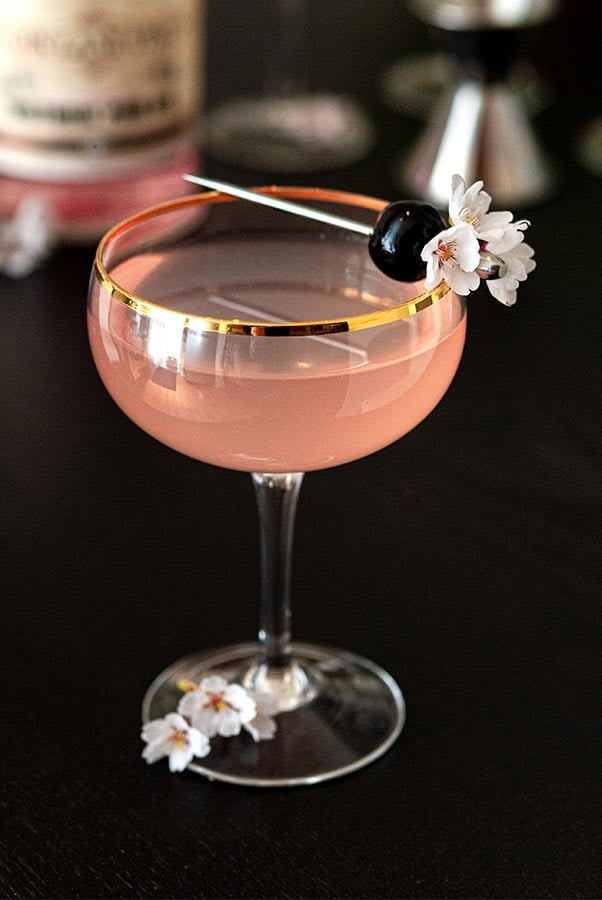 A pink cocktail, garnished with a cherry and small cherry blossoms on a black table.