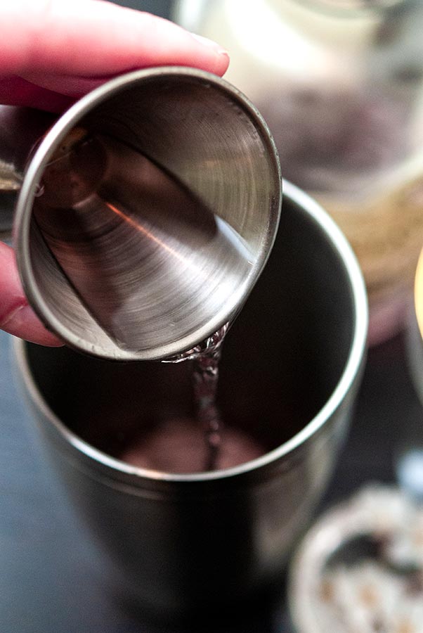 Fingers pouring maraschino liqueur from a jigger into a cocktail shaker.