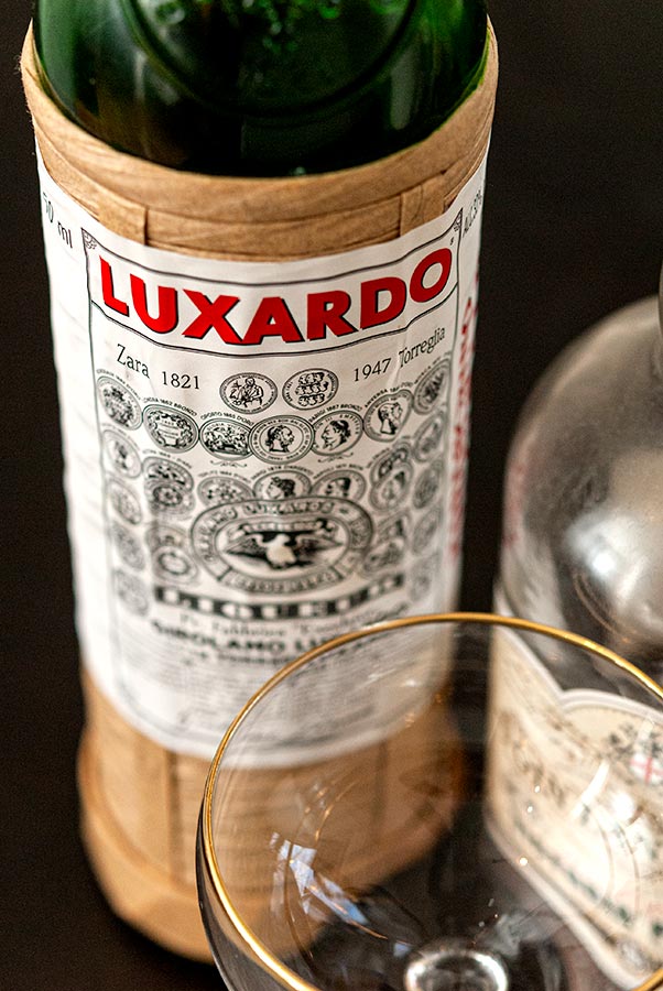 The lable on a Luxardo Maraschino Liqueur bottle, beside another bottle and cocktail glass.