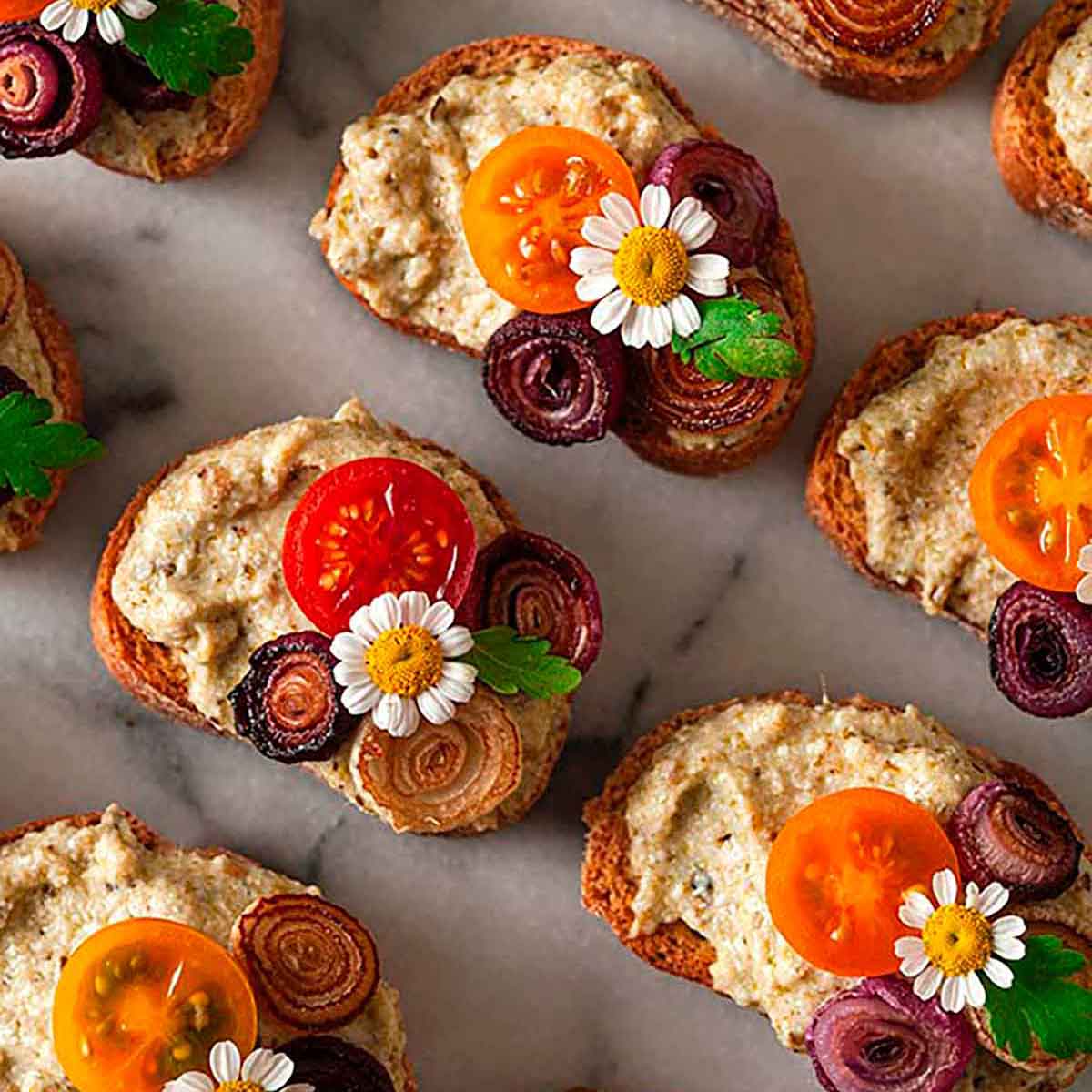 Crostini garnished with daisies, onions and tomatoes on a marble plate.