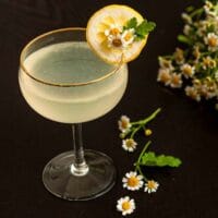 A Lemon Drop cocktail, garnished with a lemon slice and daisies on a table, with a bouquet in the background.