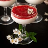 A glass of panna cotta with strawberry rhubarb compote on a table, topped with little flowers, and flowers on the table.