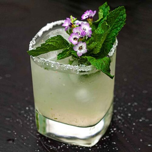 A cucumber mint margarita, garnished with small flowers and mint, on a table lightly sprinkled with salt.