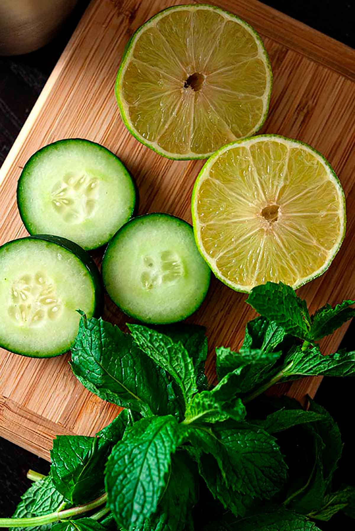 A small cutting board with 3 cucumber slices, 2 lime slices and a sprig of fresh mint.