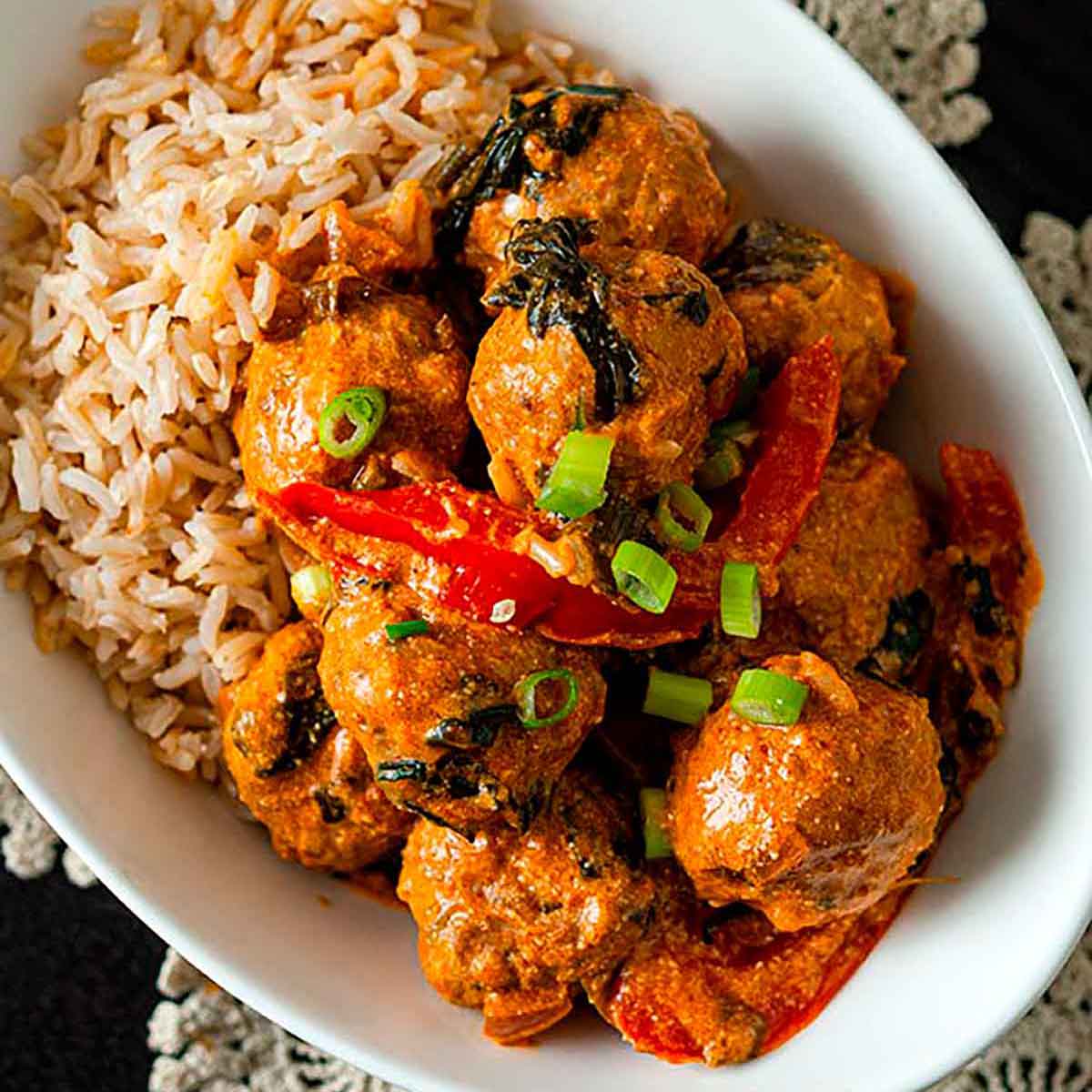 A bowl full of rice and coconut curry meatballs on a black table with a lace doily.