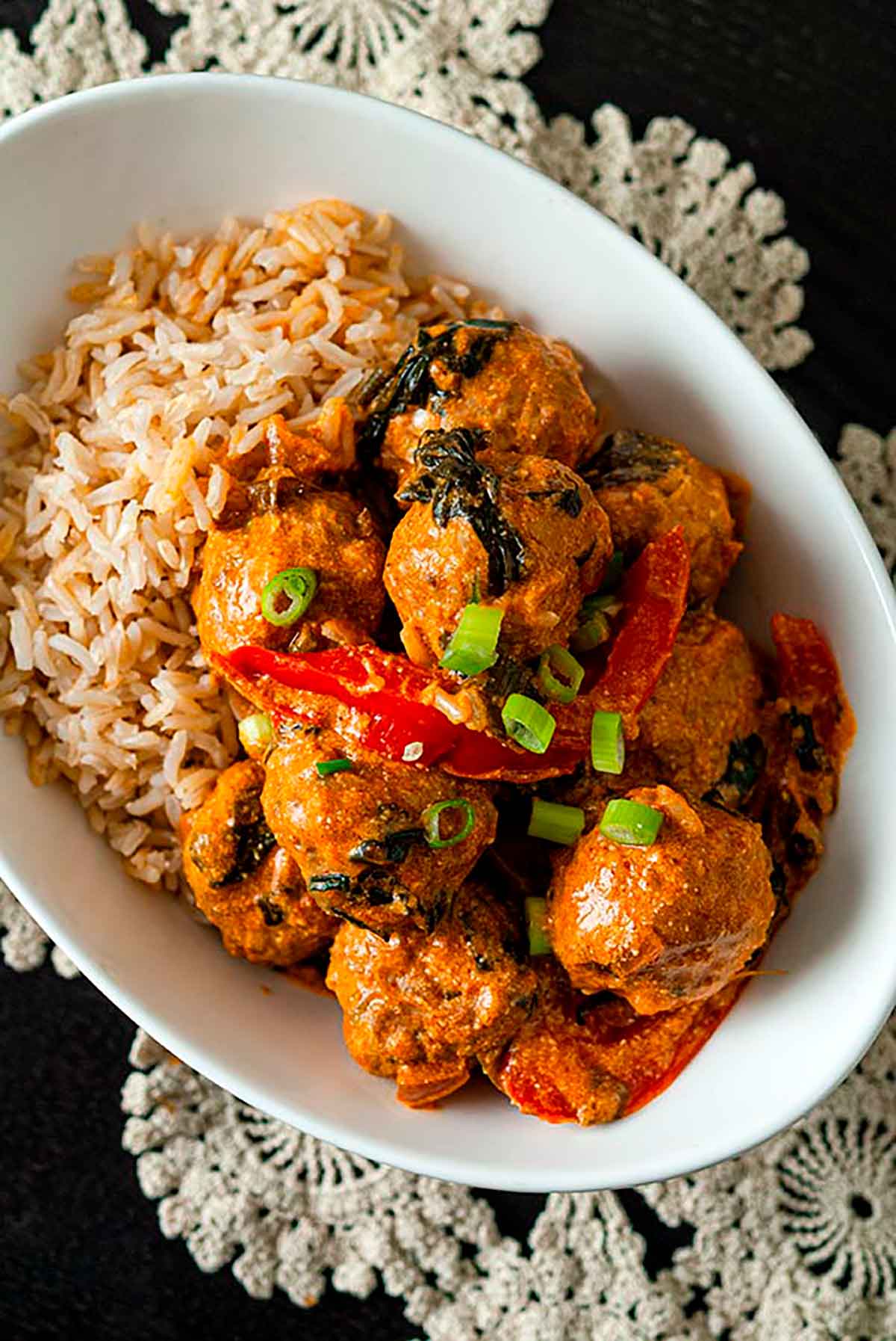 A bowl full of rice and coconut curry meatballs on a black table with a lace doily.