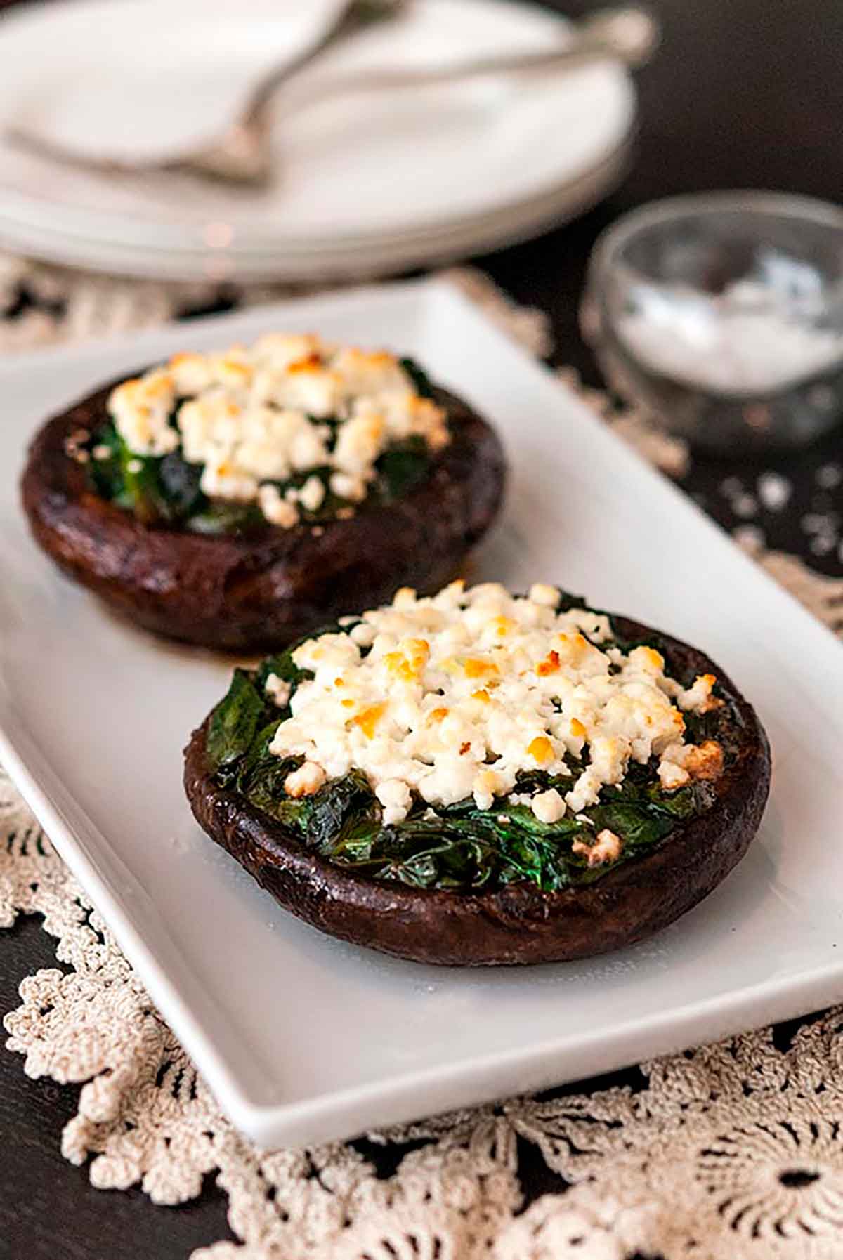 2 spinach and feta-stuffed mushroom caps on a plate on a table, lined with lace with 2 stacked plates in the background.