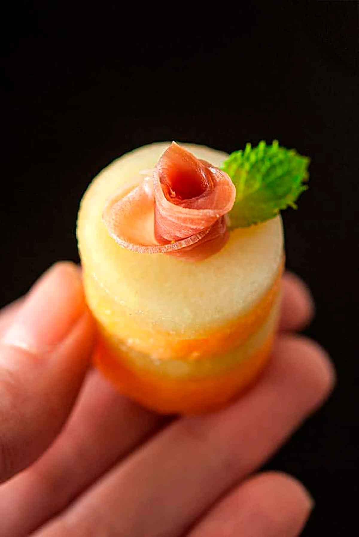 Fingers holding a prosciutto melon appetizer.