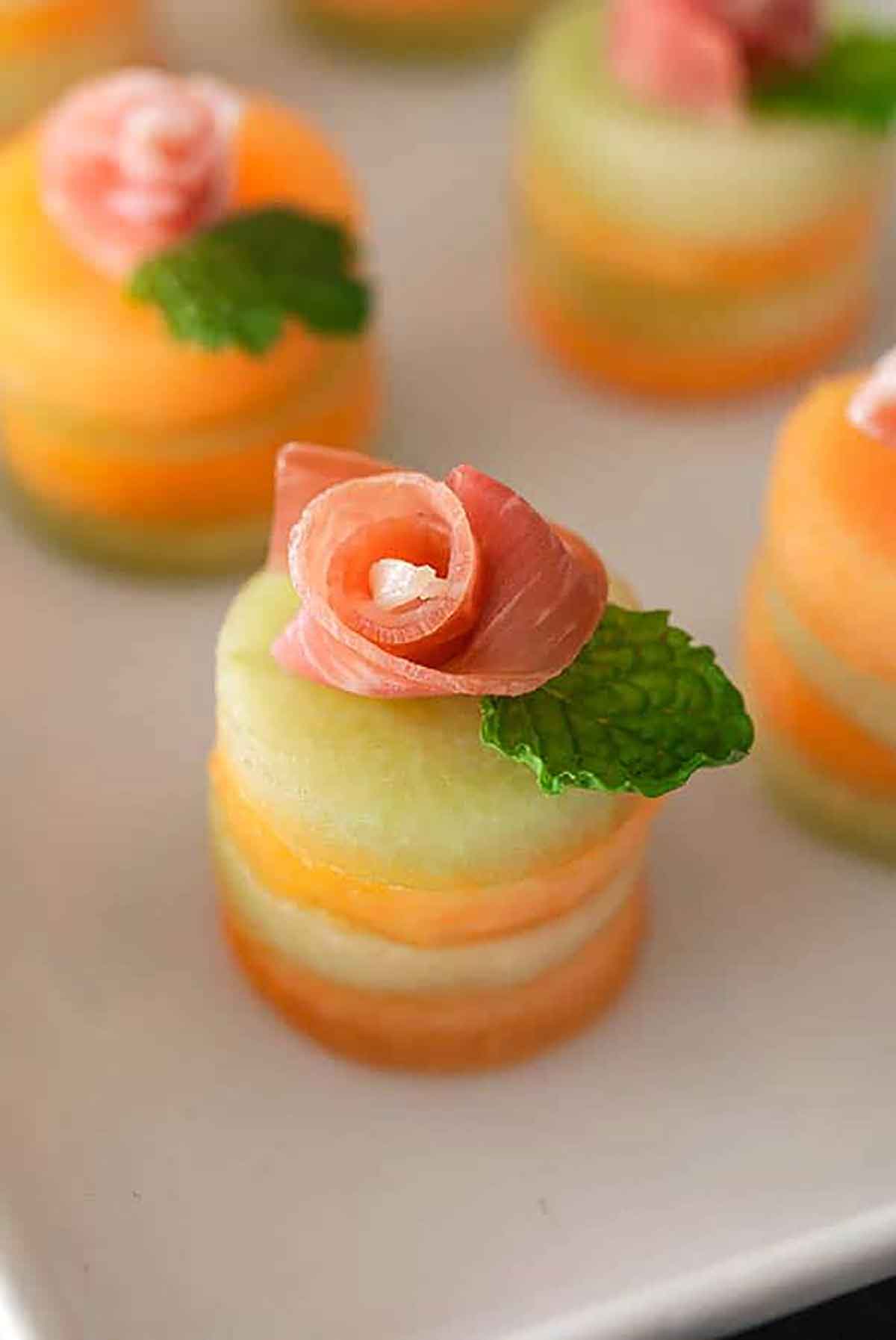 4 small melon appetizers with prosciutto roses and mint leaves on top.