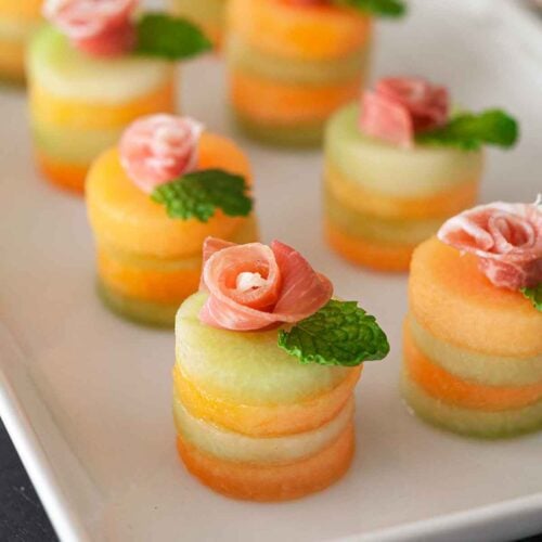 6 small melon appetizers with a prosciutto rose and mint leaves on top, in front of a lace tablecloth.