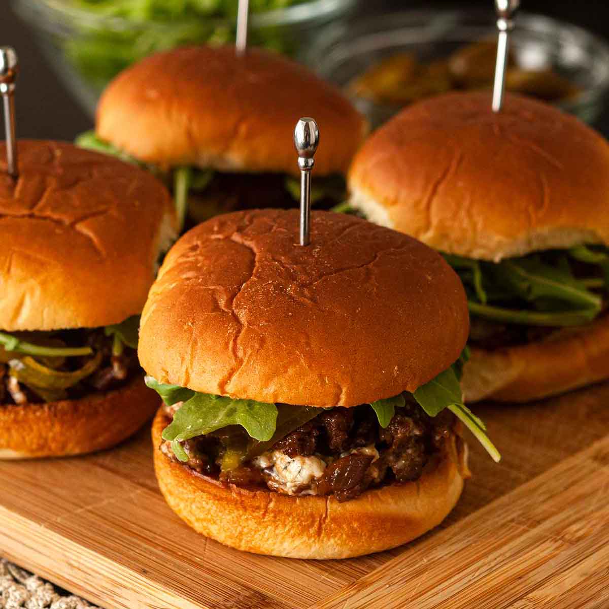 4 barbecue sliders on a cutting board above a lace table cloth with garnishes in the background.