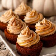 6 carrot cupcakes on a plate in front of white pumpkins.