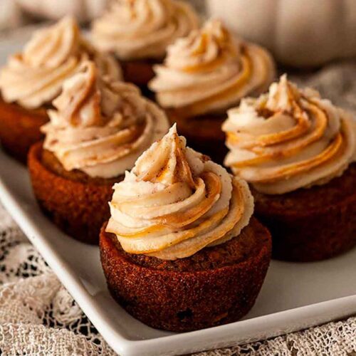 6 carrot cupcakes on a plate in front of white pumpkins.