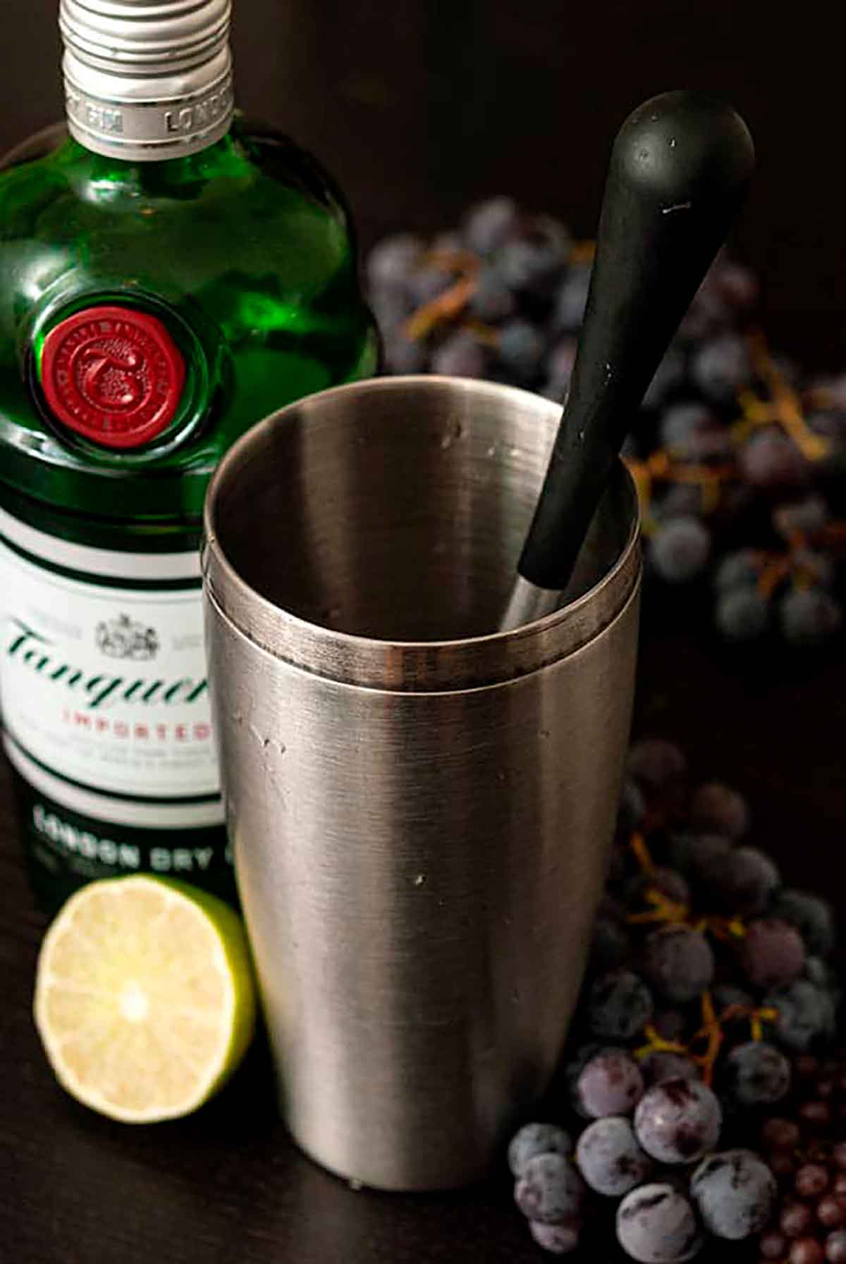 A cocktail shaker on a table with a muddler, surrounded by a bottle of gin, a lime and grapes.