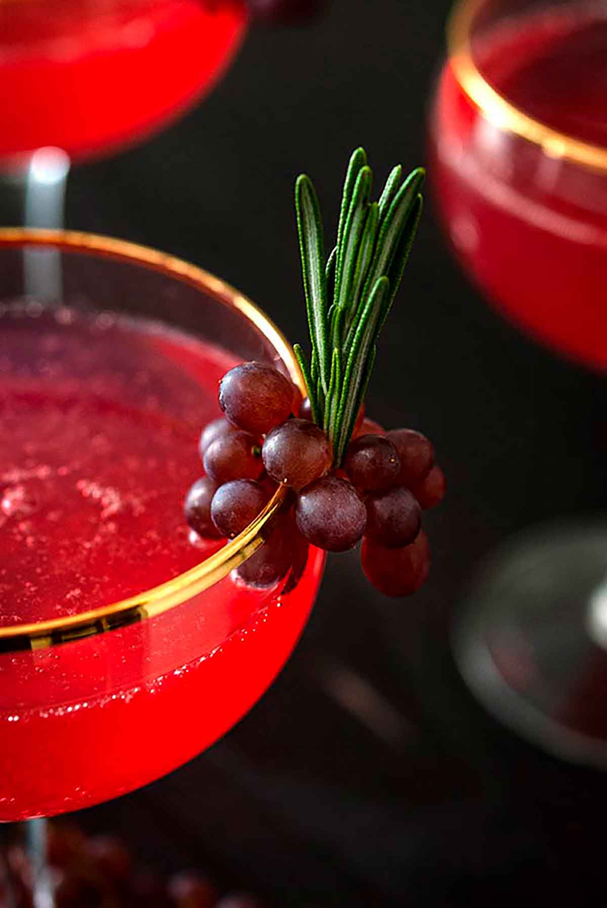 A closeup of a small grape and rosemary garnish on the edge of a cocktail glass.