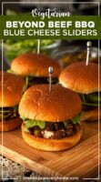 4 barbecue sliders on a wooden board on a lace tablecloth in front of bowls of garnishes.