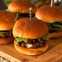 4 barbecue sliders on a wooden board on a lace tablecloth in front of bowls of garnishes.