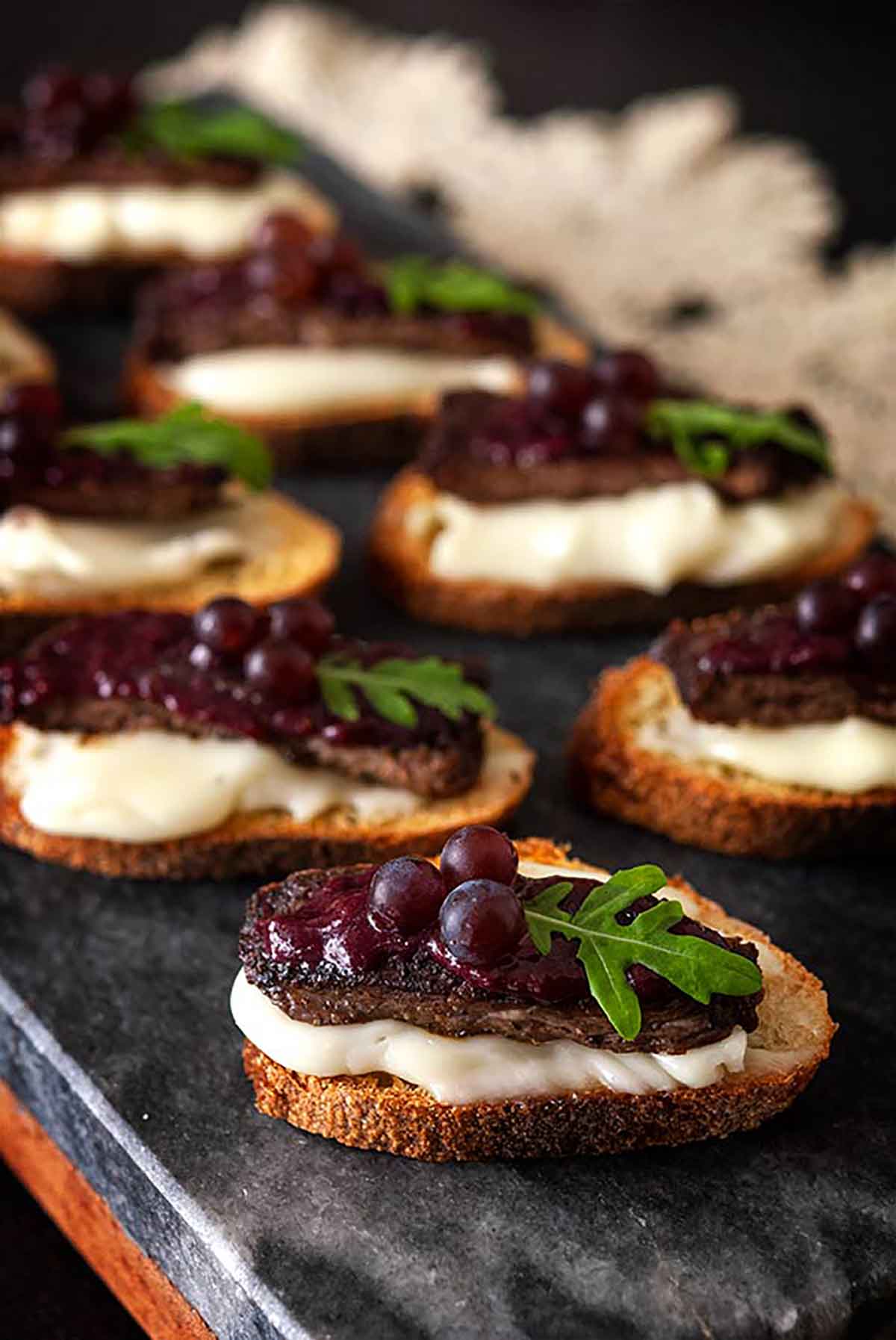 6 steak appetizers on a marble slate, garnished with tiny grapes, arugula and dripping cheese.