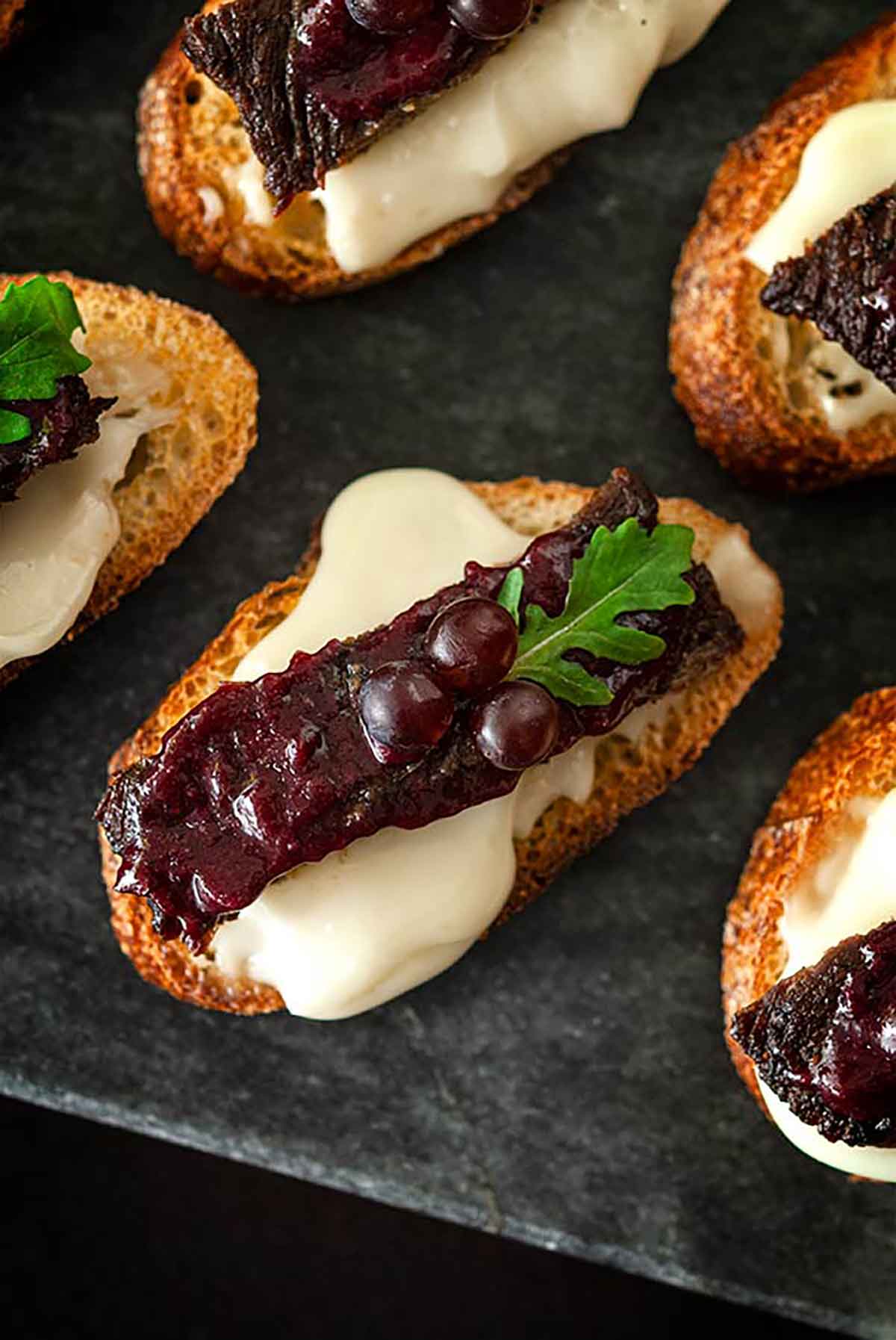 A steak appetizer, garnished with tiny grapes and a single leaf, on a marble slate, with cheese dripping over the sides.