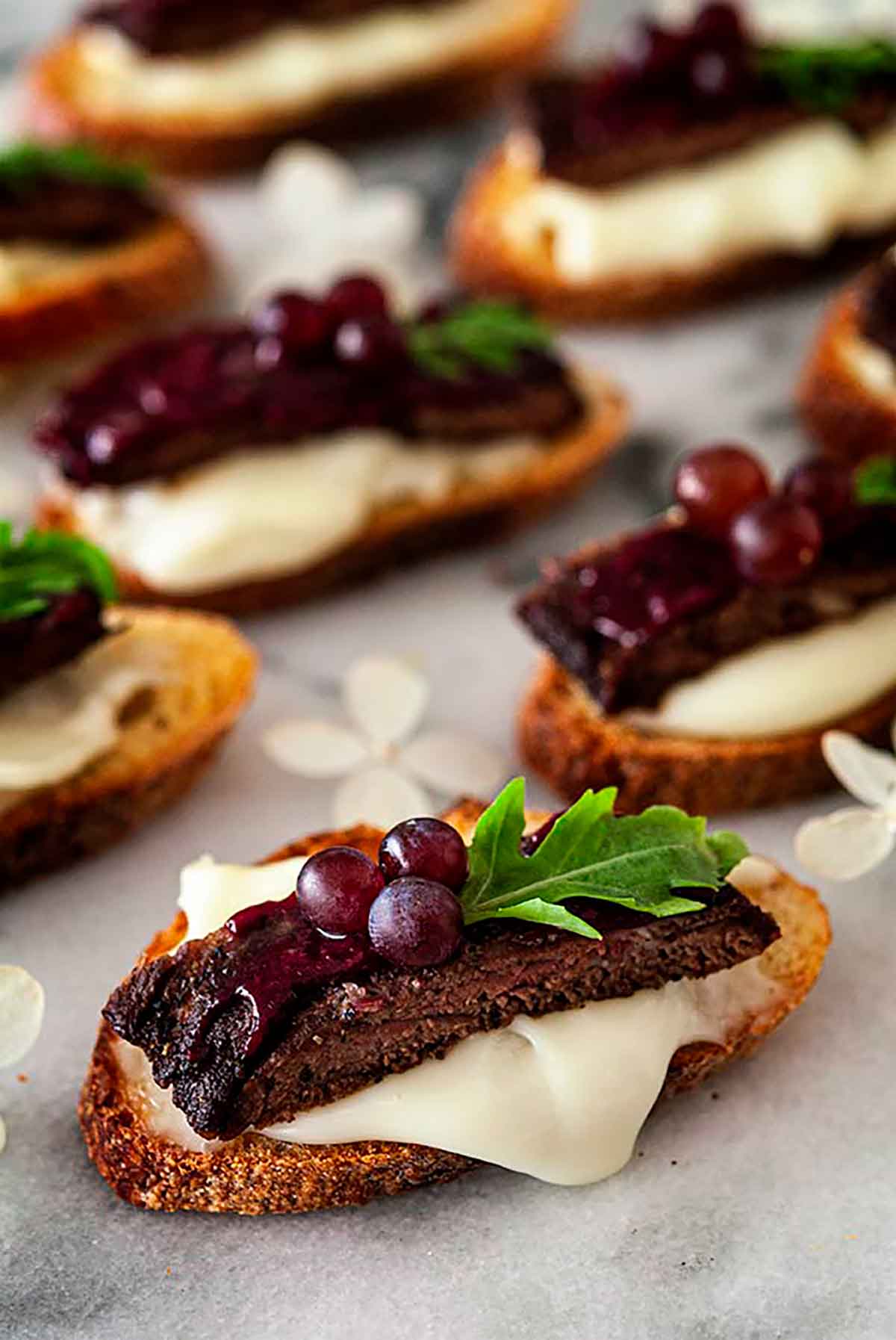7 steak appetizers on a marble slate, garnished with tiny grapes, arugula and dripping cheese.