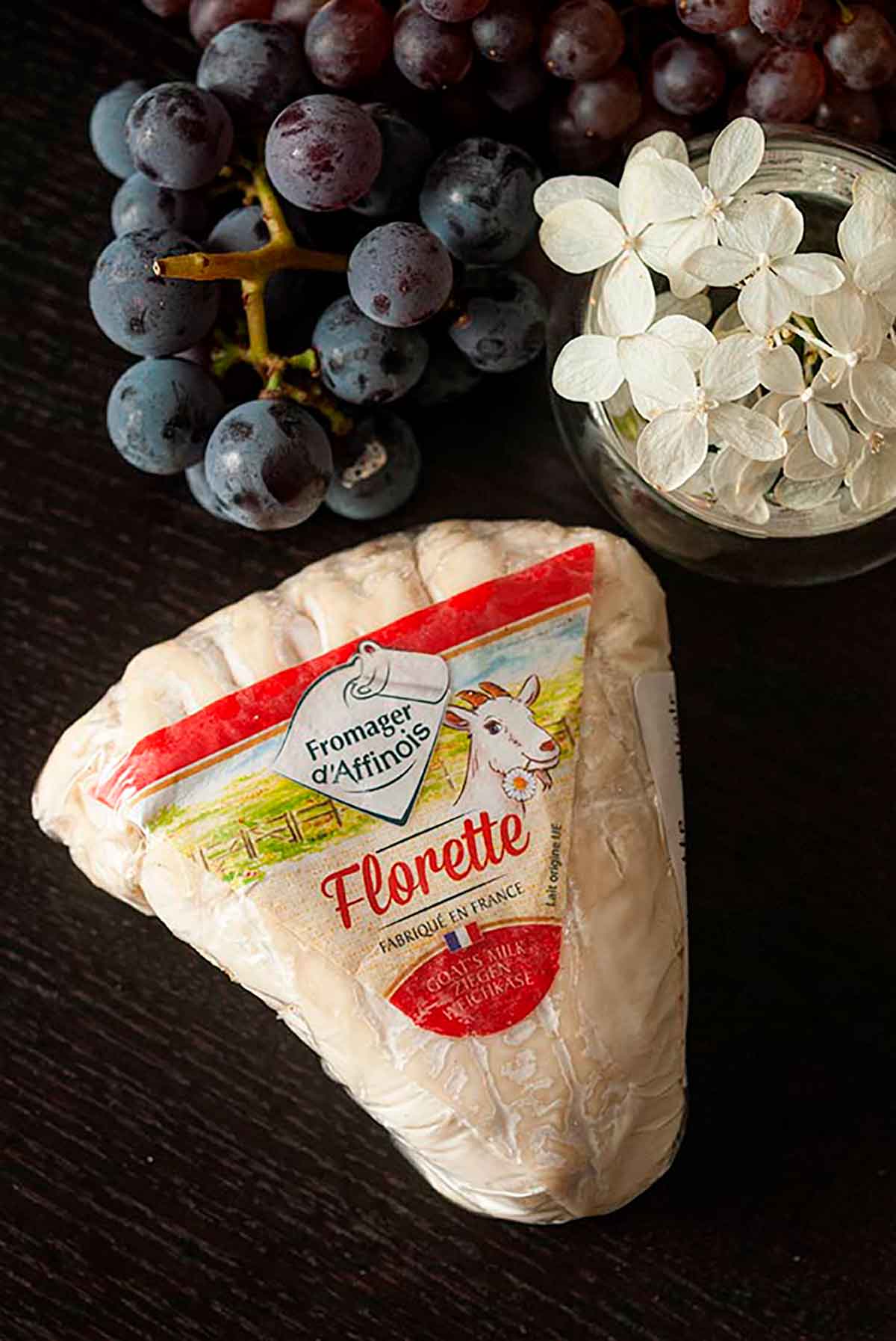 A package of Florette cheese on a table beside a small bunch of grapes and a small bowl of flowers.