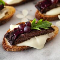 A steak appetizer, garnished with tiny grapes and a single leaf, on a marble slate, with cheese dripping over the sides.