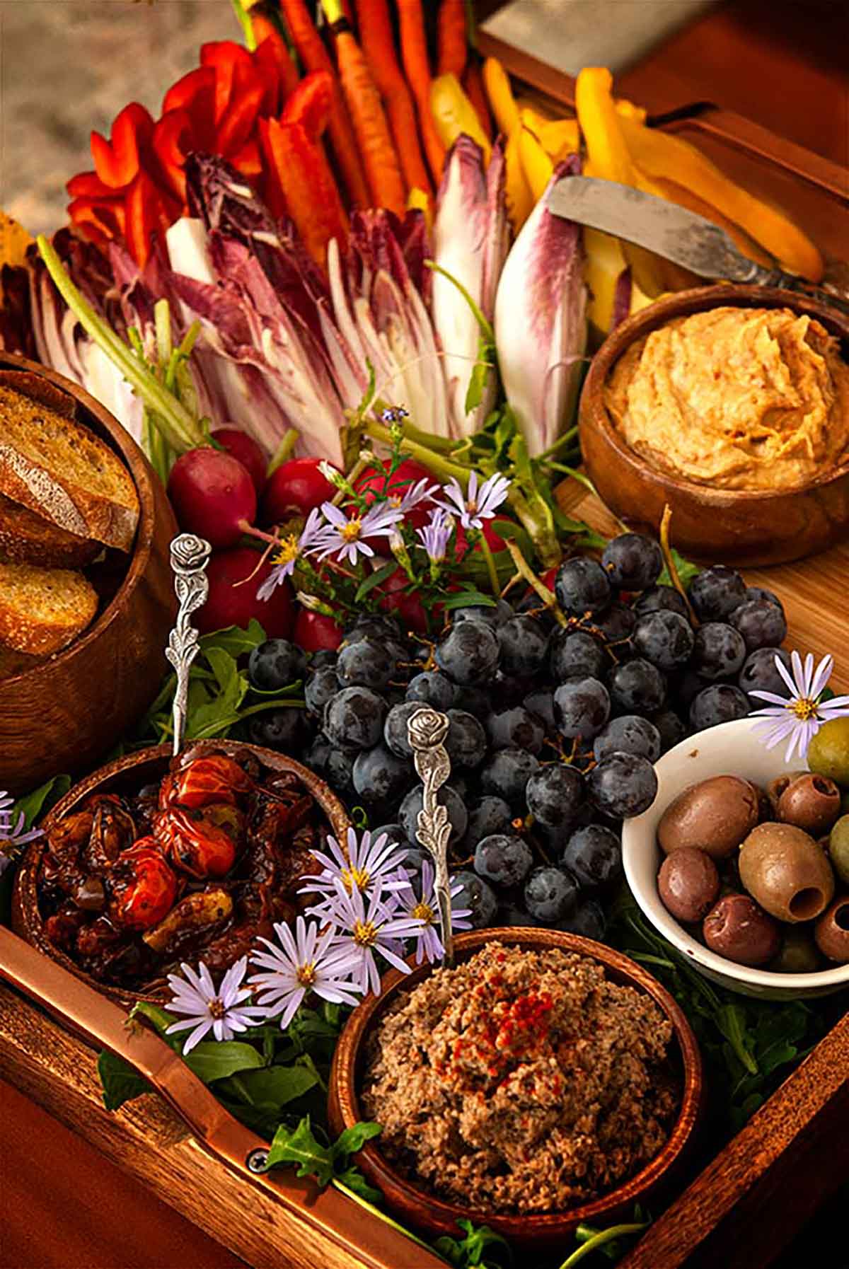 A crudités platter with vegetables, dips, olives, grapes and a bowl of bread on a tray.