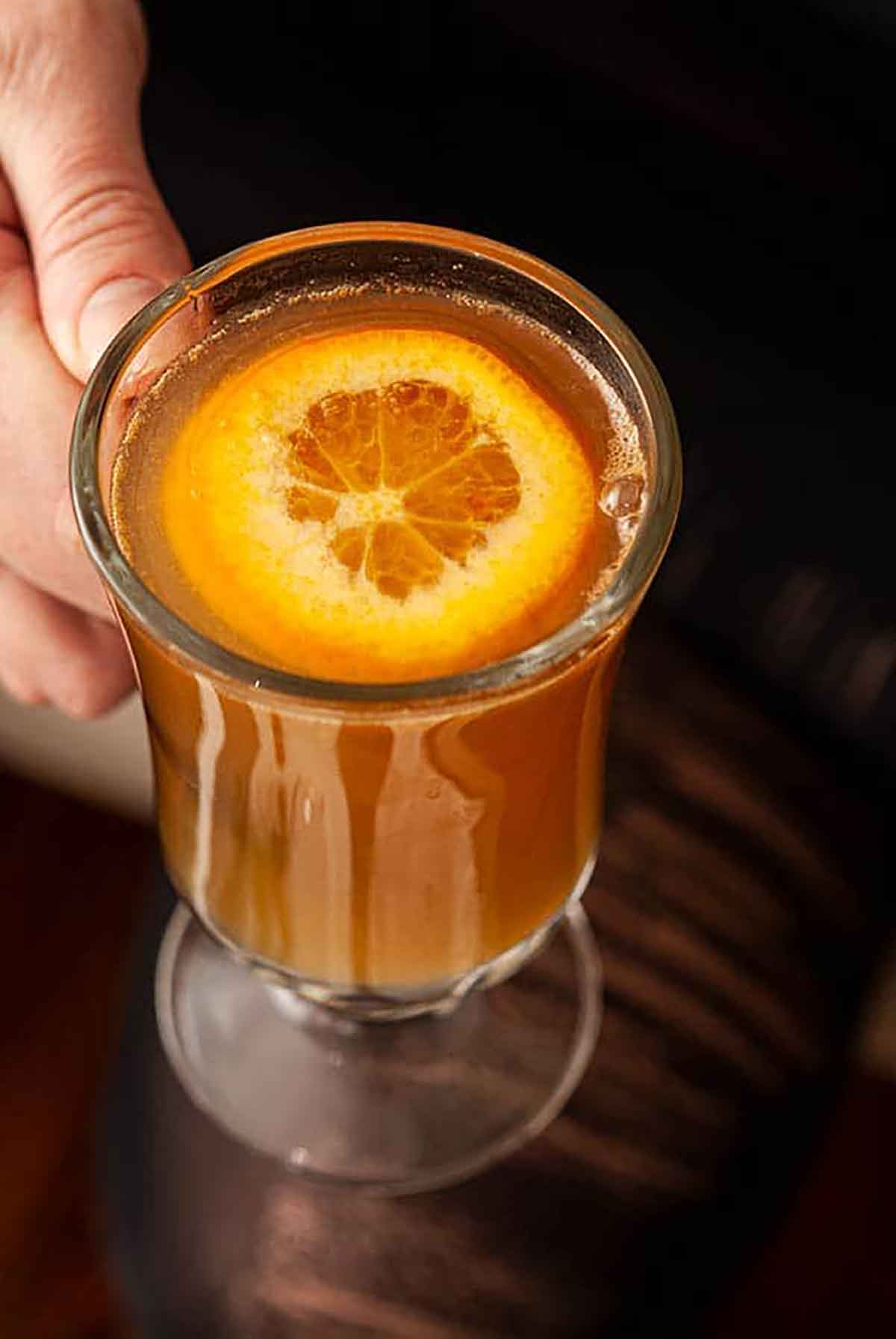 A hand holding a mug of cider whisky, garnished with a sliced orange.