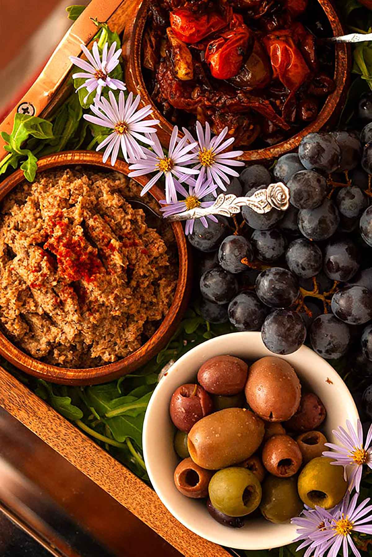 Bowls of bruschetta, pate and olives surrounded by grapes, arugula and flowers in a tray.