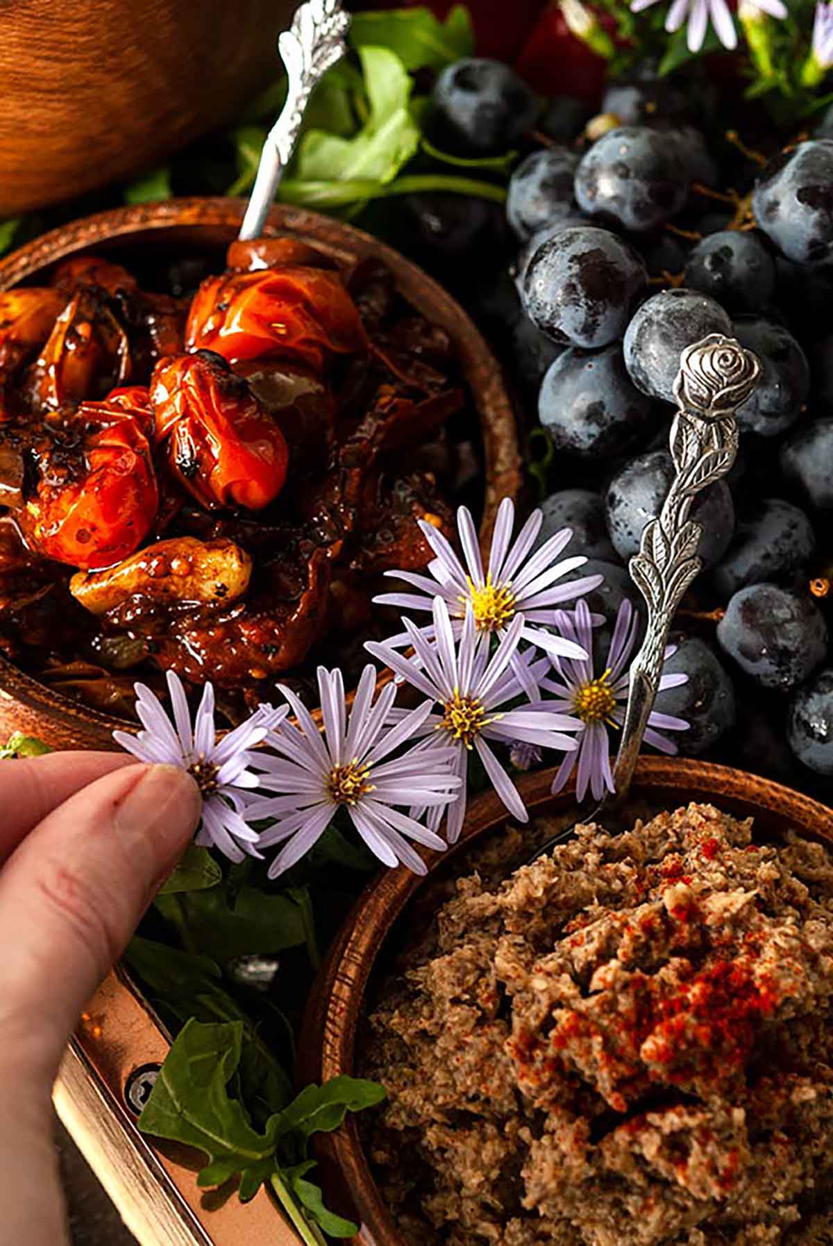 Fingers adding flowers between bowls of dips, surrounded by grapes and arugula in a tray.