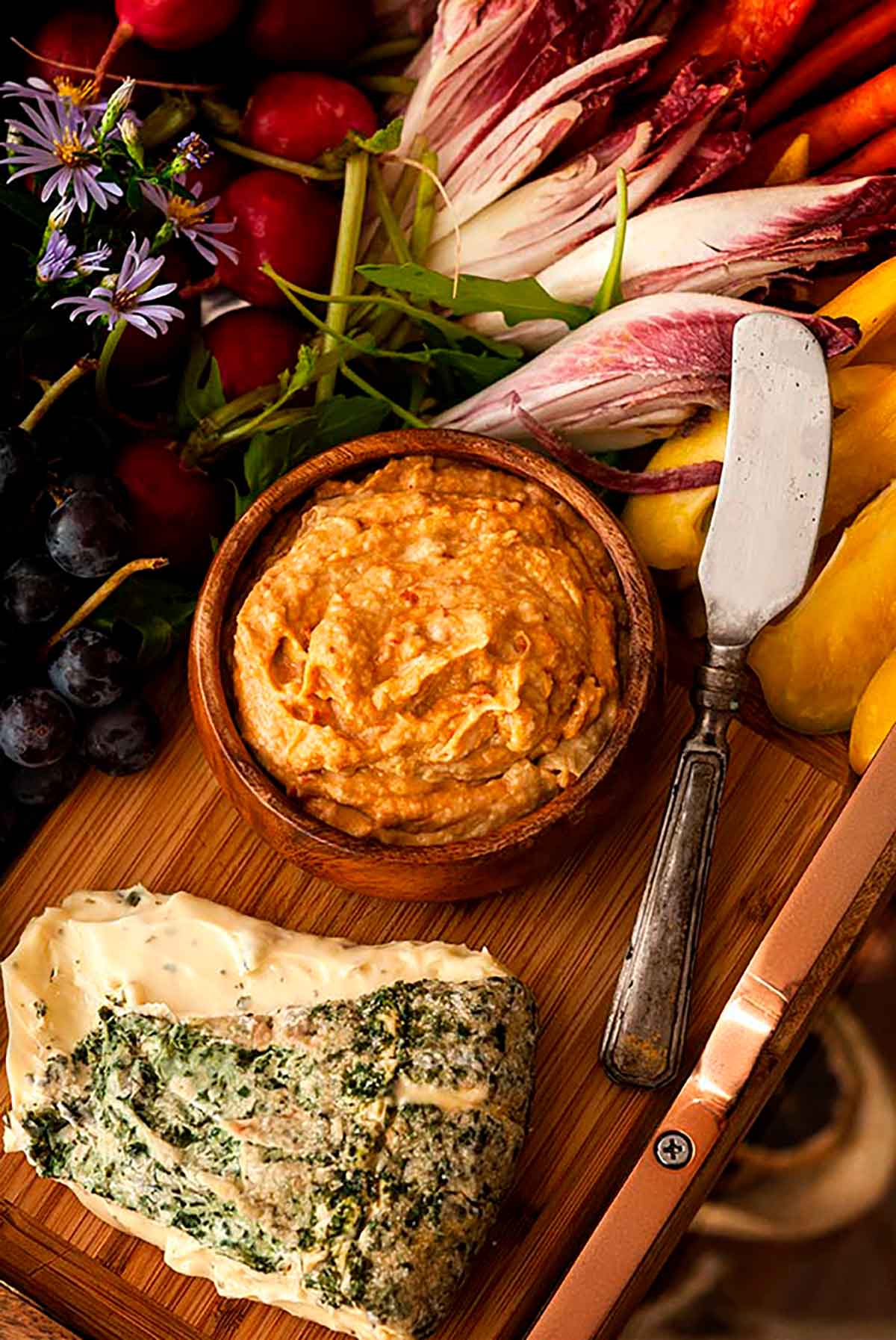 A bowl of hummus on a board with herbed cheese and a cheese knife, surrounded by vegetables.