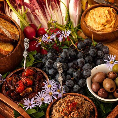 A crudités platter with vegetables, dips, olives, grapes and a bowl of bread on a tray.