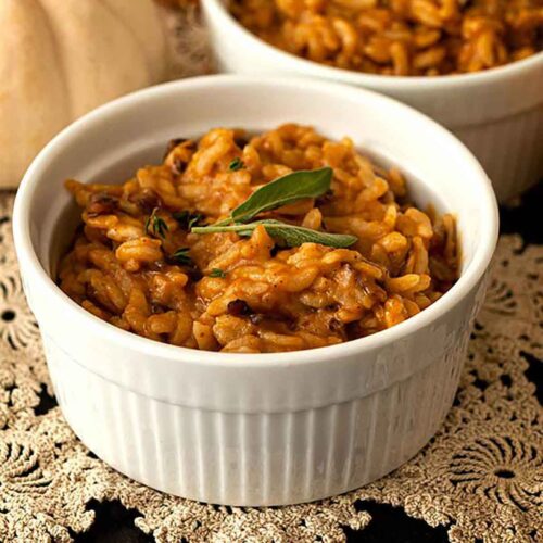 2 bowls of pumpkin risotto, garnished with sage leaves, on a lace table cloth with 3 small pumpkins in the background.