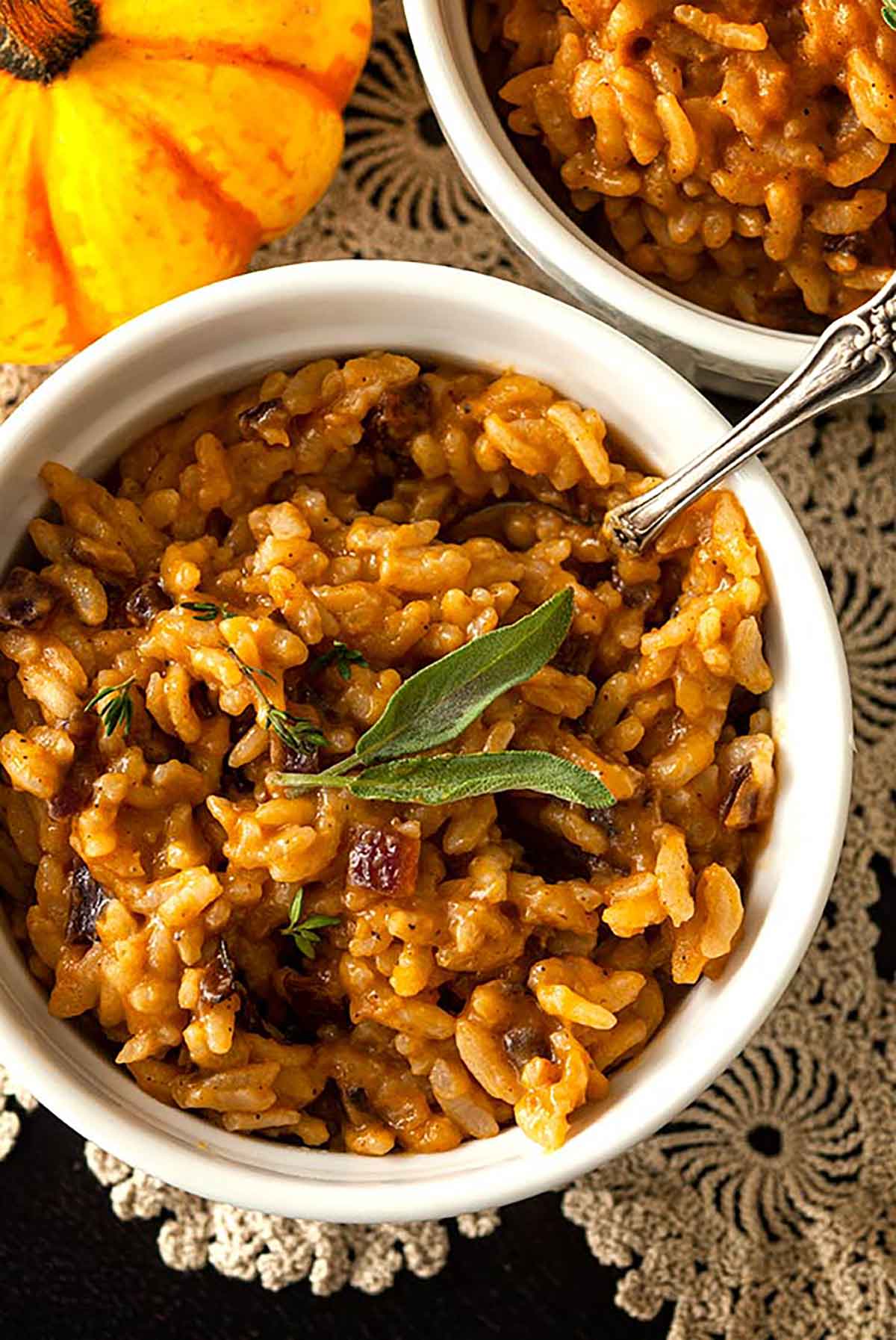 A bowl of pumpkin risotto, garnished with 2 sage leaves, on a lace table cloth beside another bowl and small pumpkin.